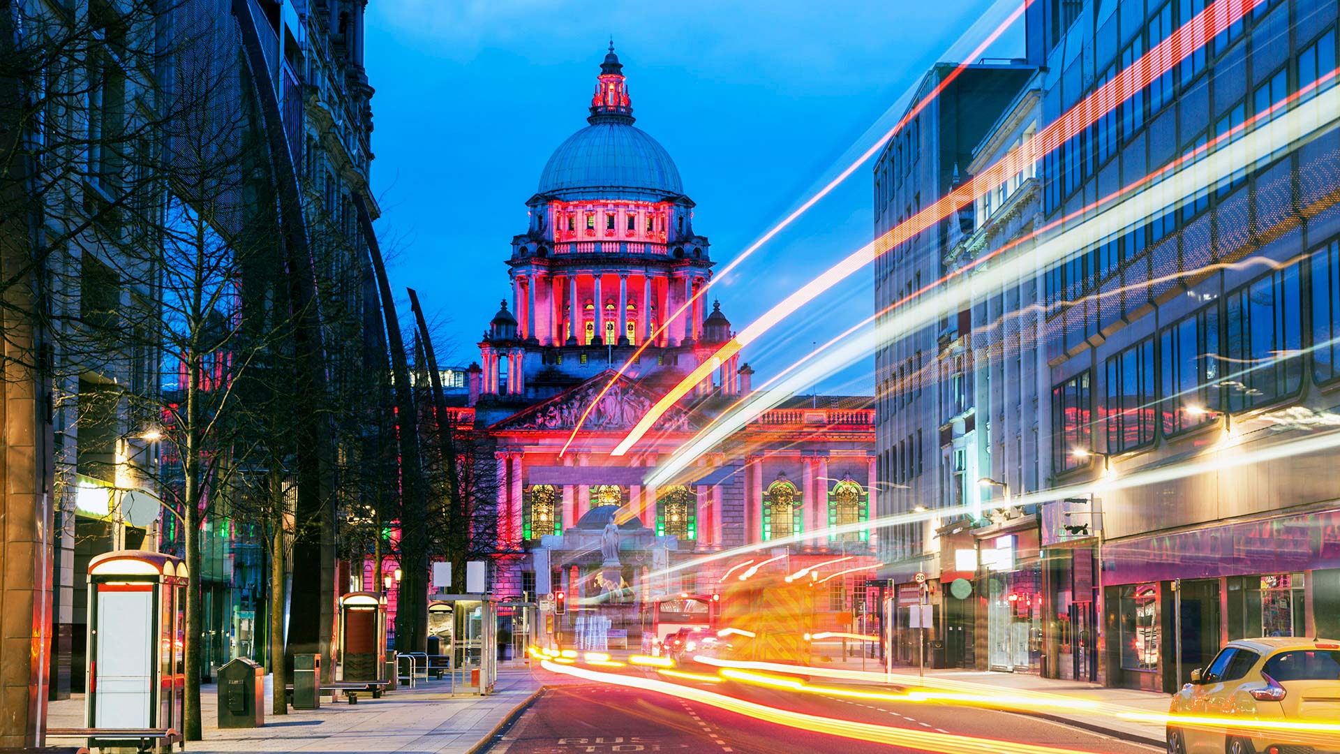 Belfast City Hall illuminated at night with colorful light trails from passing traffic in the city centre.