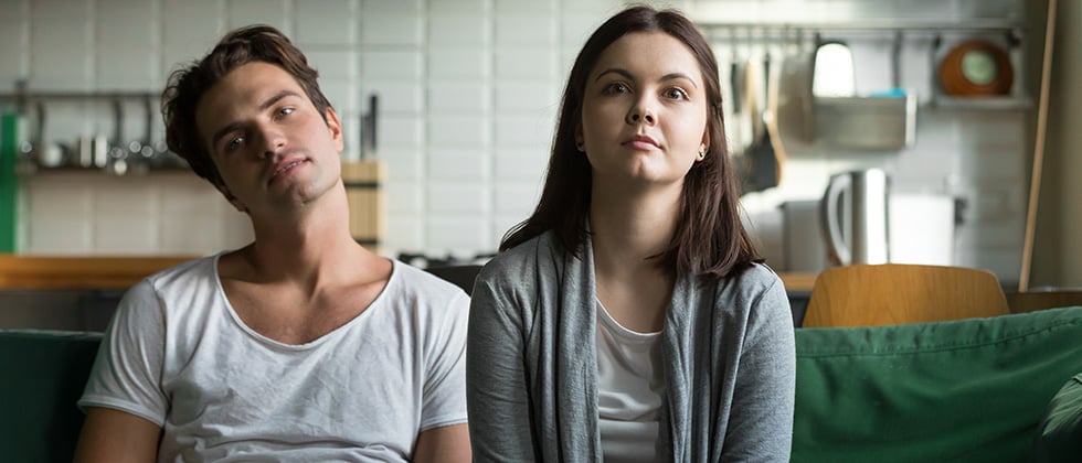 Bored-looking couple sit together on a sofa, staring forward vacantly