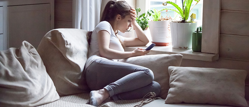A woman sits alone on a living room sofa, looking at her phone