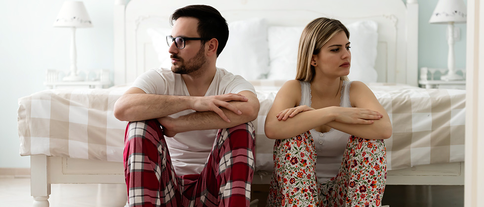 A couple in pajamas sit at the end of their bed, looking away from each other