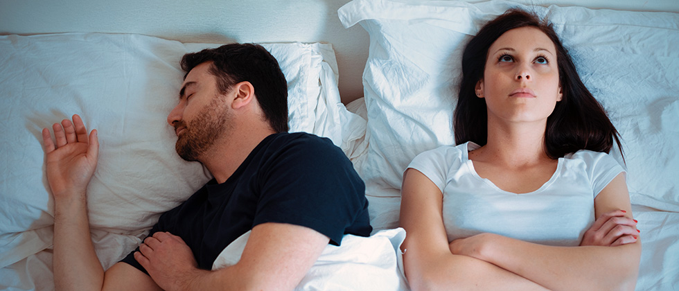 A couple in bed, the man asleep and the woman looking at the ceiling, bored