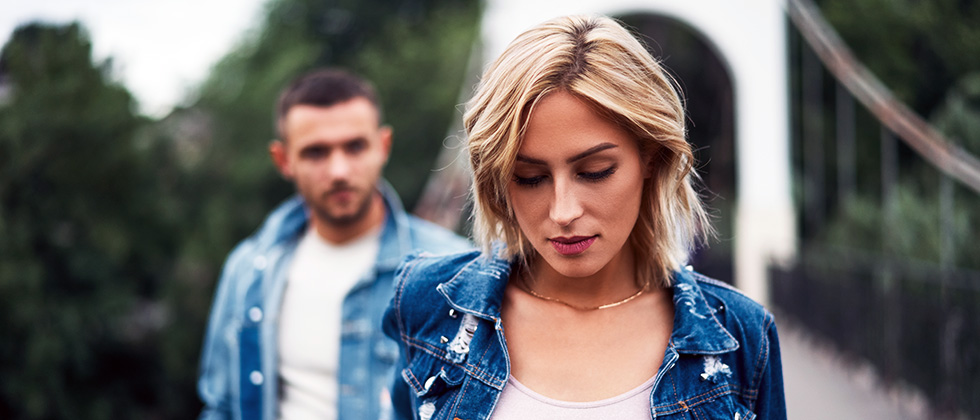 A woman waks in front of her partner, both with downcast expressions