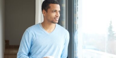 A young guy staring out a window holding coffee and smiling