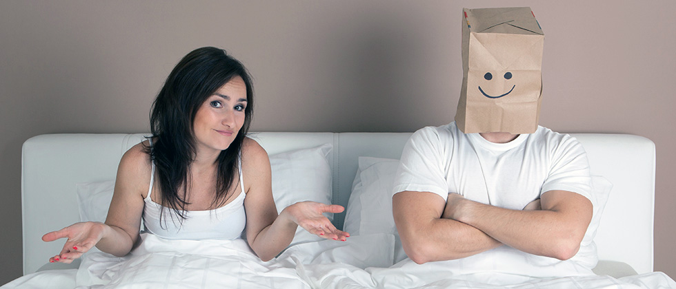 A couple in bed, the woman shrugs her shoulders, the man wears a paper bag over his head