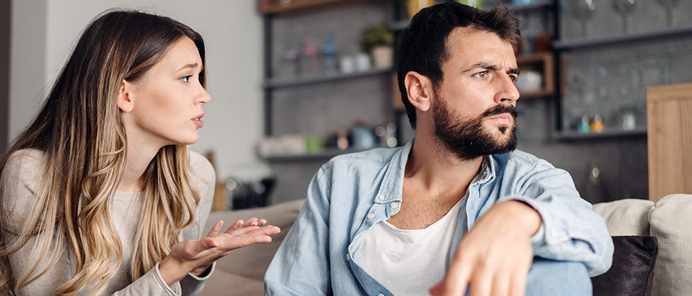 A couple sit on a sofa, the woman making a pleading gesture towards the man as he looks away