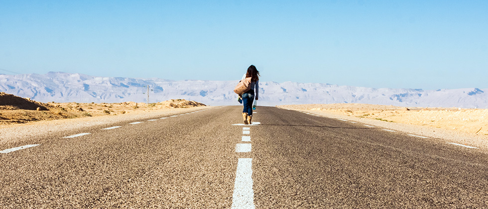 A woman walks away down an empty road in a desert with mountains on the horizon