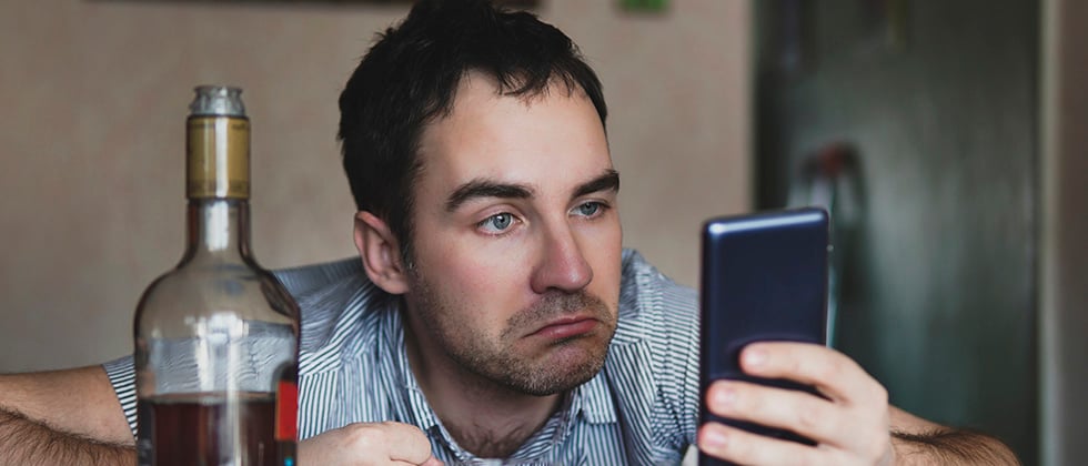 A drunk-looking man stares at his phone, a half-empty bottle of spirits in front of him