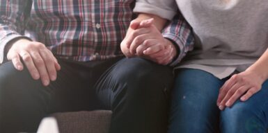 A couple sitting other with their hands held together resting in their lap