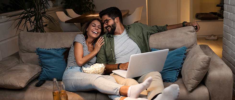 Woman and a men sitting on the sofa with popcorn watching a movie on the laptop and having date night at home