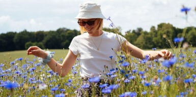 A woman standing on a field of violet flowers. She looks happy and is waring a hat and sunglasses.