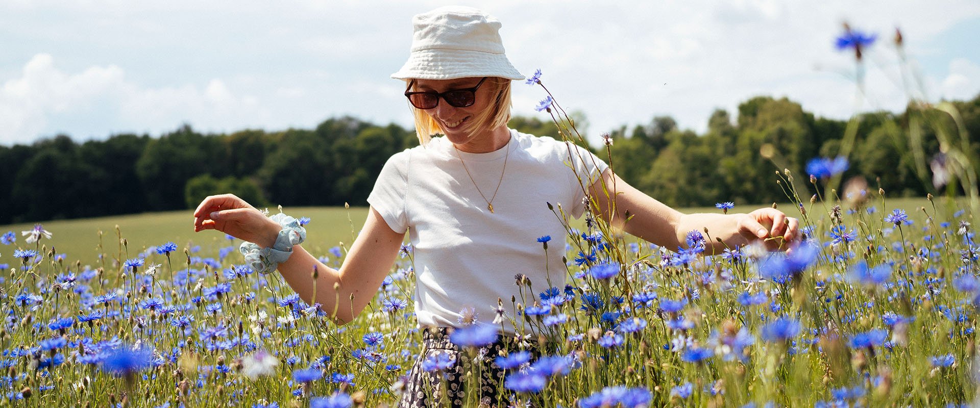 A woman standing on a field of violet flowers. She looks happy and is waring a hat and sunglasses.