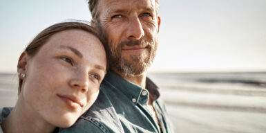 A couple is on the beach and the woman is leaning her head on the mans' shoulder. The woman seems to be younger than the man.