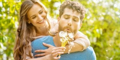 A woman on her date's back holding dandelions while he blows on them