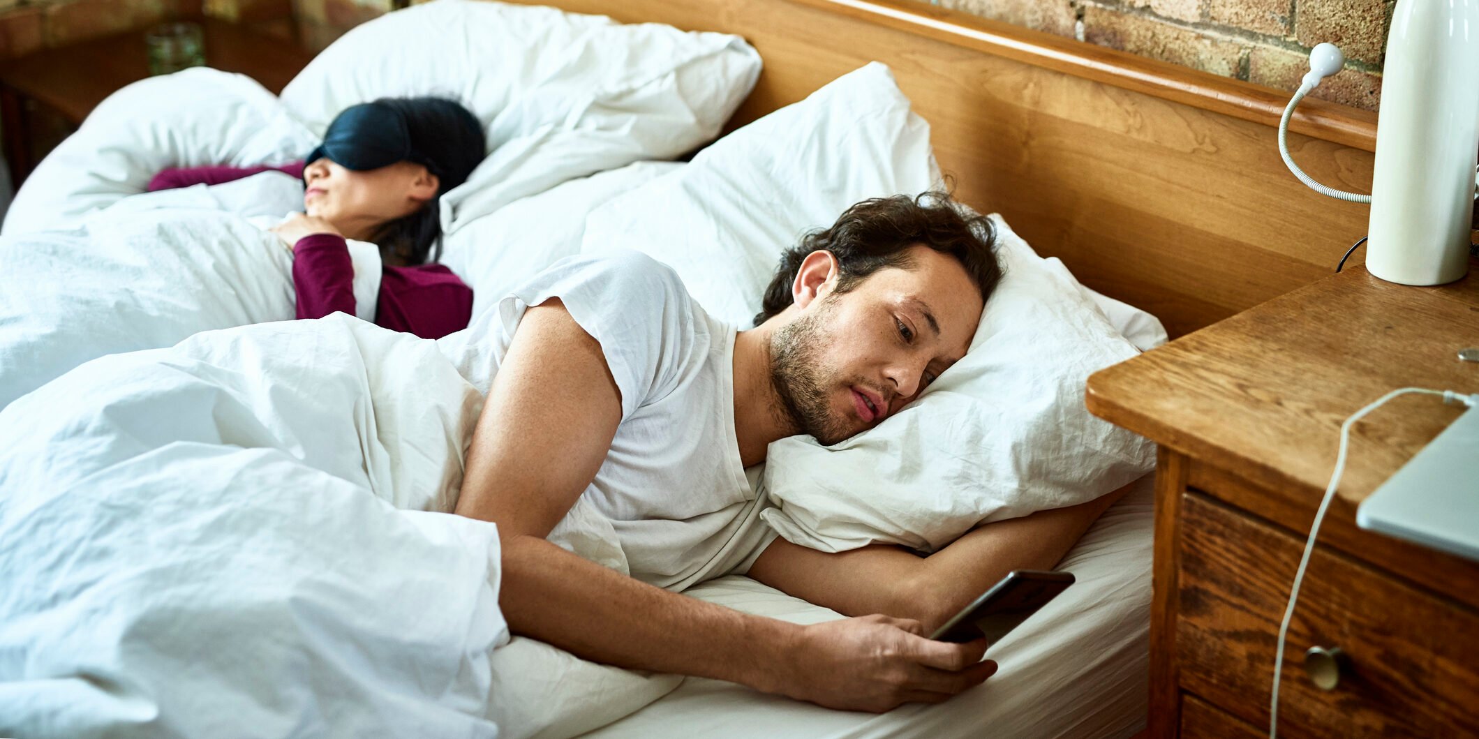 Woman fast asleep next to partner who is checking his smartphone