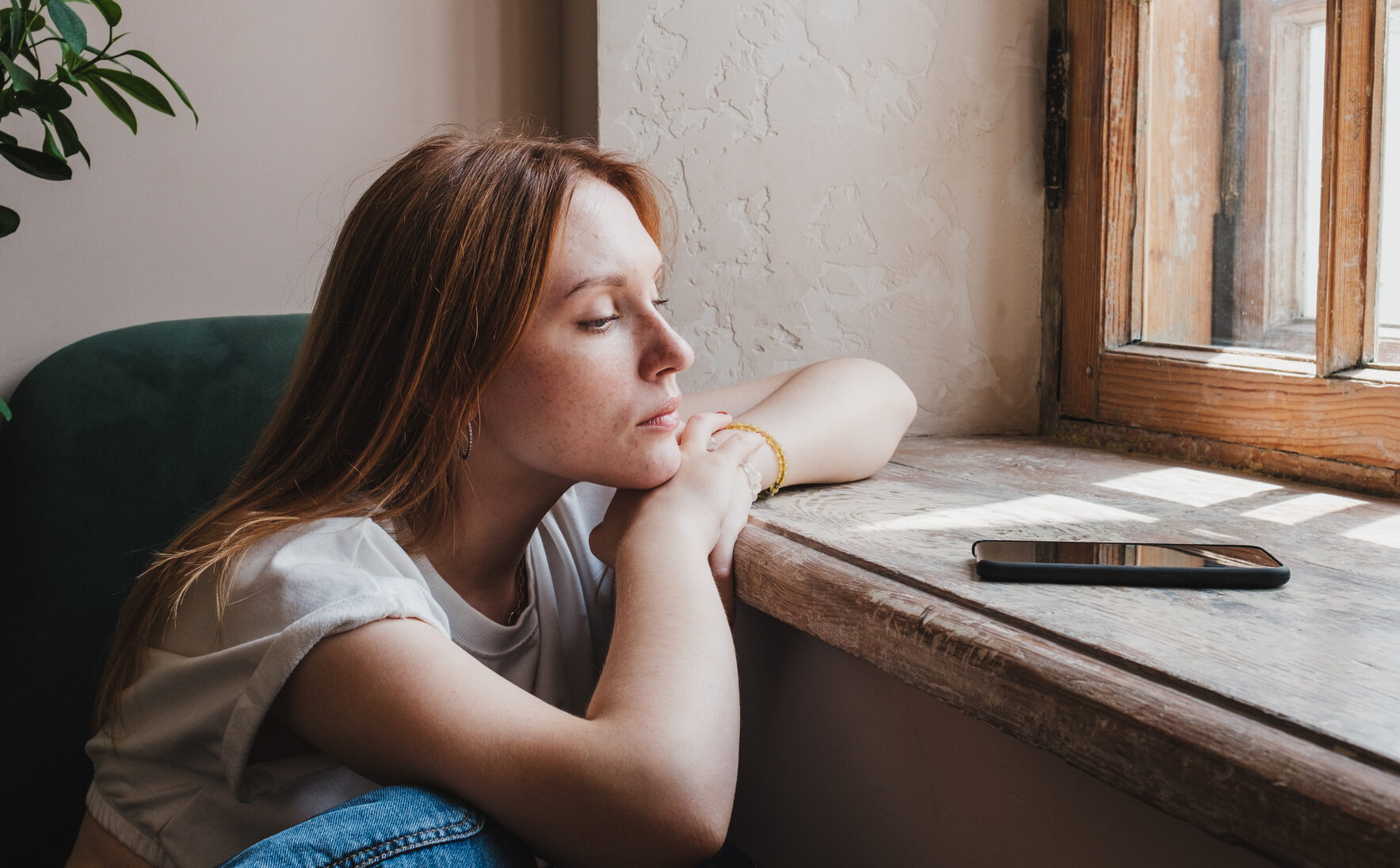 Upset redhead teen girl sitting by window looking at phone waiting call or message