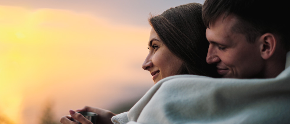 A couple cuddled under a blanket looking at the sunset outside