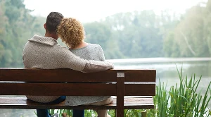 A man is holding a woman in his arms and they are sitting at a park bench with river views.