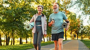 A man and a woman are walking together in a park, each carrying a yoga mat under their arm.