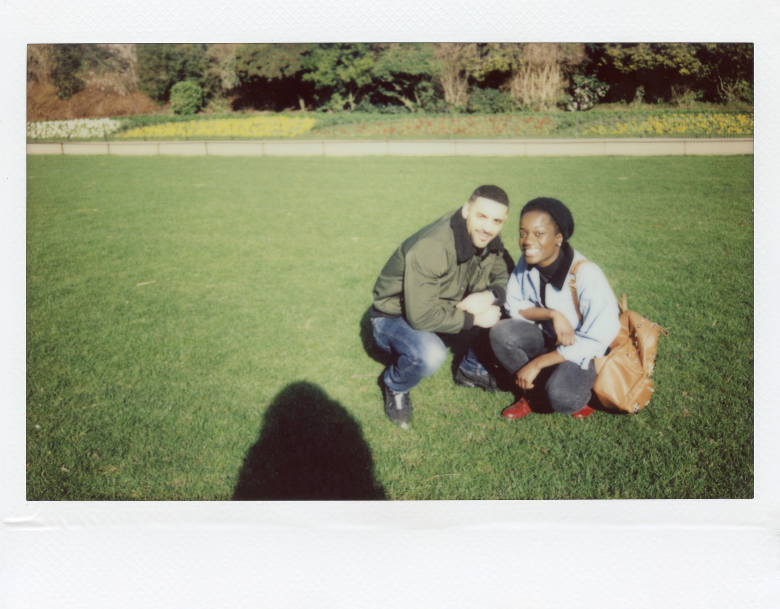A polaroid-style grainy photo with a man and woman wearing denim pants and jackets sitting in a grassy park smiling at the camera.