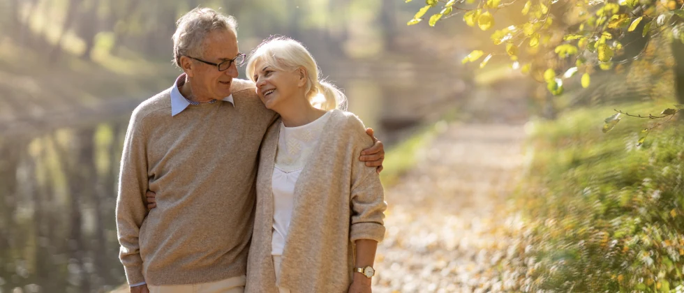 A couple walk side by side along a gravel path near a forest in the sunlight.