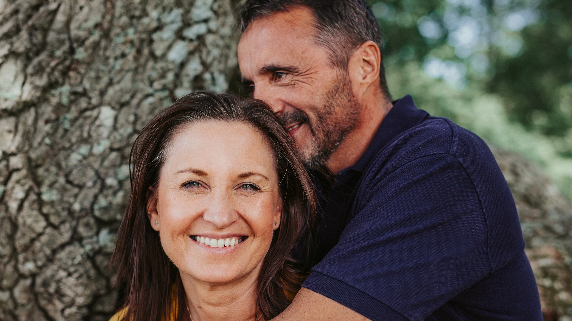 A man is hugging a woman in front of a tree.