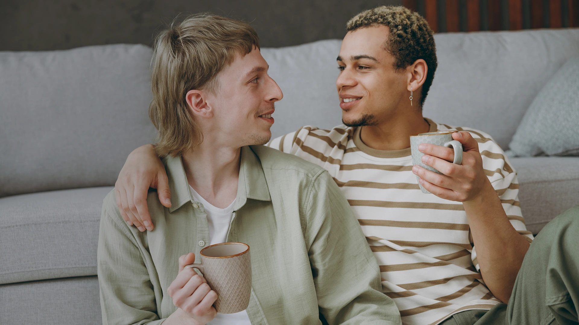 A gay couple are enjoying a cup of coffee together on their sofa.